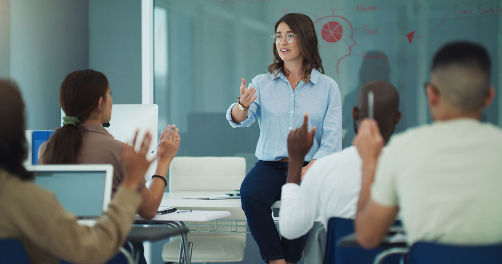 A teacher standing in front of a class calls on a student within several who have their hands up.