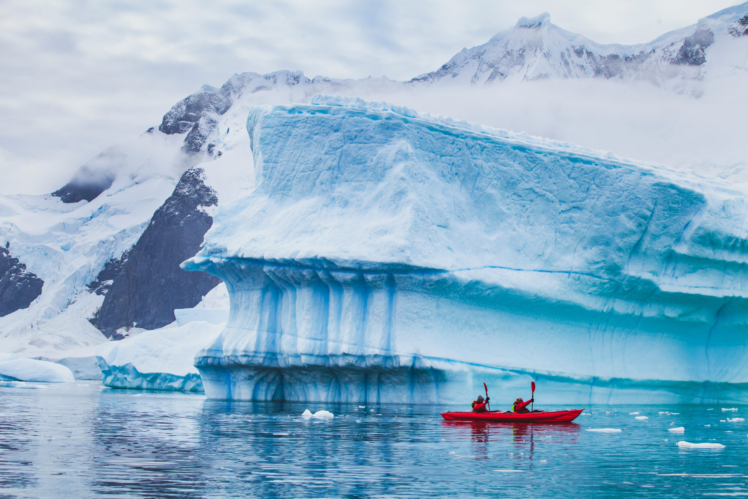 A pair of kayakers navigate past an iceberg near a rocky Antarctic coast
