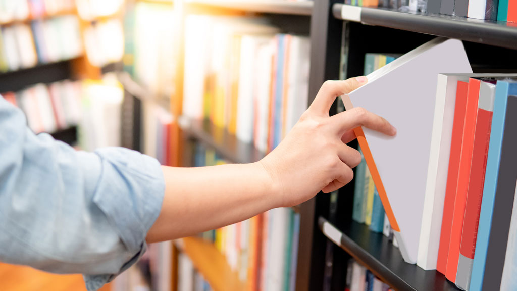 Arm-length view of hand pulling a book from a library shelf full of books