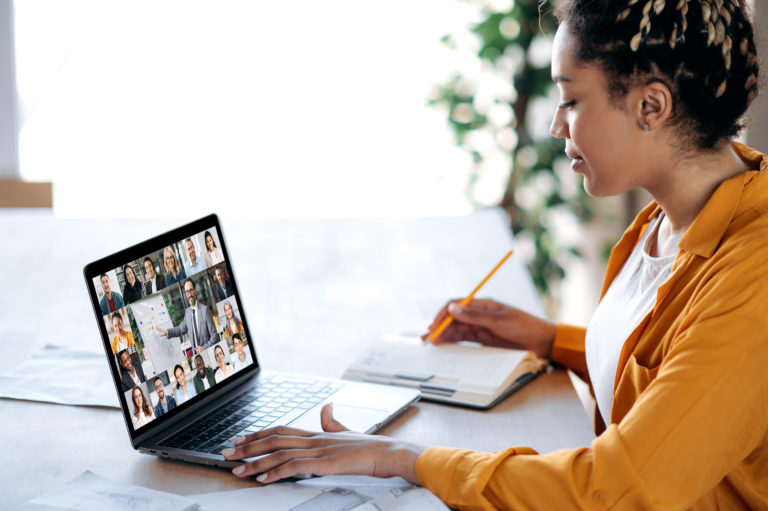 Photo of African American woman participating in an online program on her laptop while taking notes