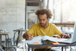 Photo of African American student writing notes in notebook with book and laptop at a cafe table
