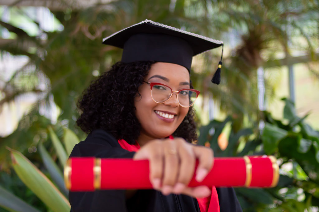 Photo of a female graduate wearing cap and gown holding out a rolled up diploma