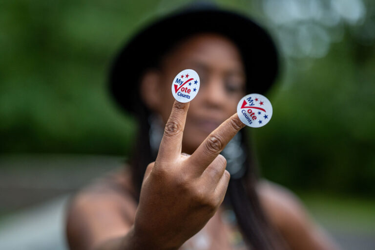 Photo of woman showing "My vote counts" sticker in foreground, while she's visible in background