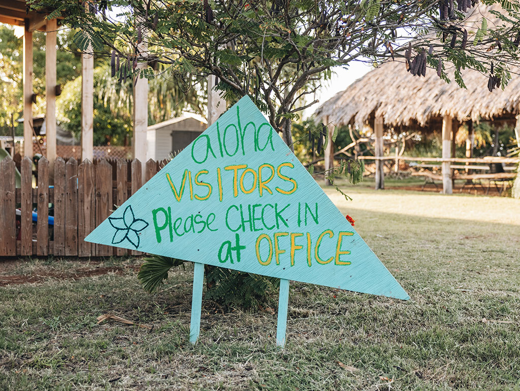 Green triangular sign saying "Aloha visitors, please check in at office"