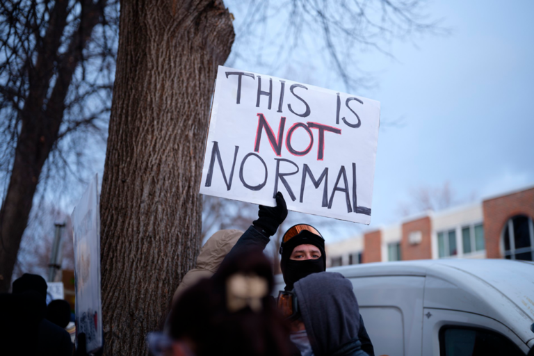 A protester in heavy winter clothing holds up a sign with the message, "This is not normal" in Minneapolis, Minn., in January 2026; © David Bowman
